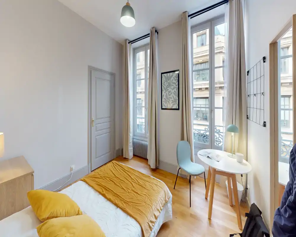 Sunny, stylish bedroom with a double bed dressed in warm linens, high windows with long curtains and a small desk by the window—bright and cozy space ideal for relaxing or working.