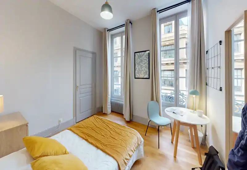 Chambre lumineuse et soignée avec lit double, grandes fenêtres habillées de longs rideaux et petit bureau près de la fenêtre — espace chaleureux idéal pour se détendre ou travailler.