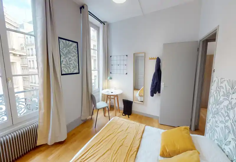 Chambre lumineuse et bien mise en scène avec un lit double aux draps jaunes, de grandes fenêtres laissant entrer beaucoup de lumière naturelle, une petite table avec chaise et un joli parquet en bois.