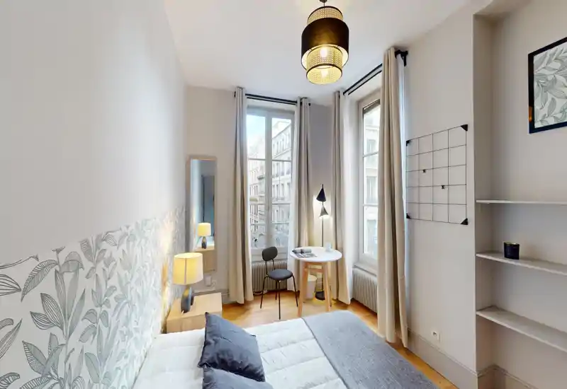 Bright, stylish bedroom with a comfortable double bed, patterned headboard, bedside table lamp and a small workspace by tall windows letting in abundant natural light.