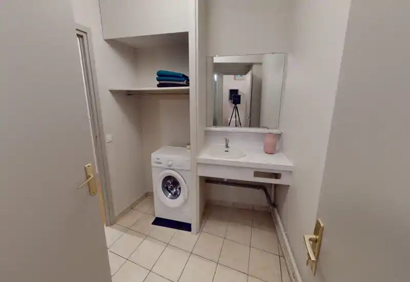 Petit espace d'entrée/lingerie avec machine à laver, lavabo et miroir ; carrelage et murs neutres pour un espace propre et fonctionnel, adapté au linge.