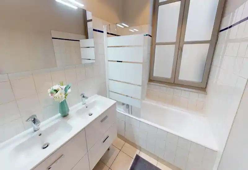 Salle de bains lumineuse et soignée avec double vasque, baignoire équipée d'une paroi en verre dépoli et fenêtre givrée apportant de la lumière naturelle.