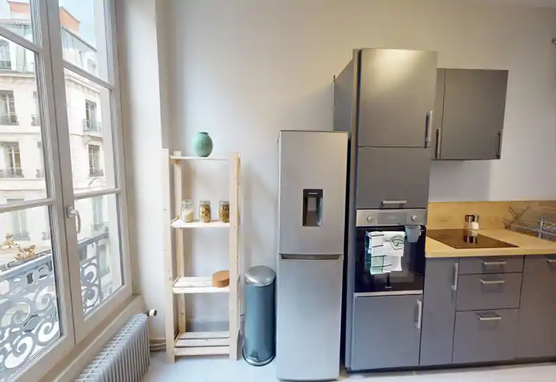 Coin cuisine moderne et lumineux avec réfrigérateur en acier inoxydable, four encastré, plaque à induction et plan de travail en bois ; une grande fenêtre apporte beaucoup de lumière naturelle et une étagère offre du rangement.