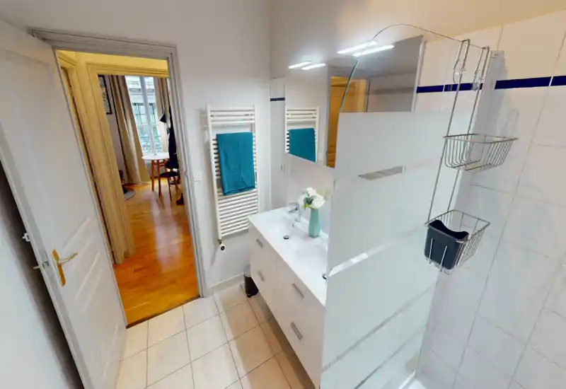 Salle de bains lumineuse et propre avec meuble vasque double, miroir éclairé et douche à paroi en verre sablé ; carrelage clair et sèche-serviettes apportent une touche moderne.