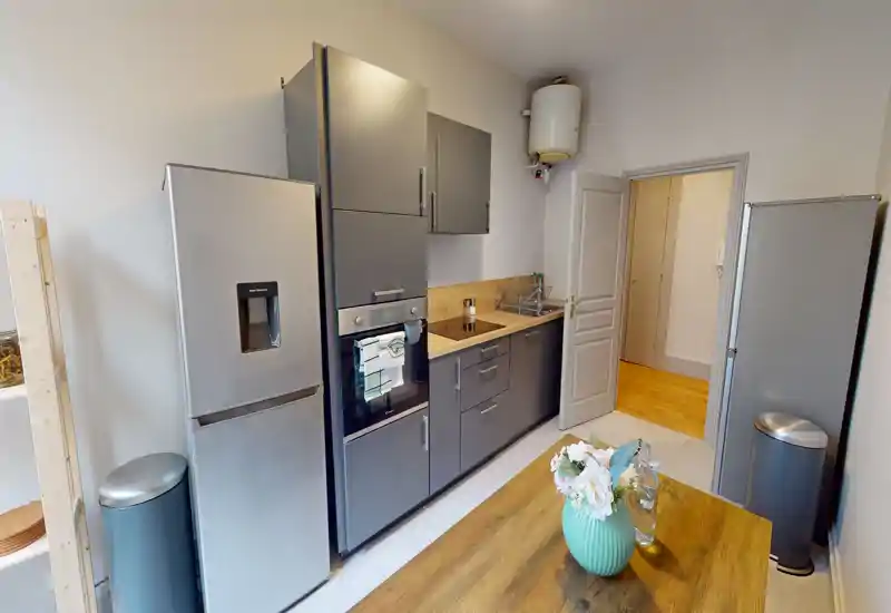 Cuisine moderne en longueur avec placards gris, four encastré, réfrigérateur en acier inoxydable et table en bois avec vase décoratif — lumineuse et bien rangée.