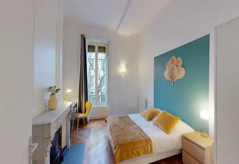 Camera da letto luminosa e di stile con parete accentata turchese, letto matrimoniale con biancheria ocra, comodino con lampada e una piccola scrivania vicino alla finestra — parquet e camino decorativo donano carattere.