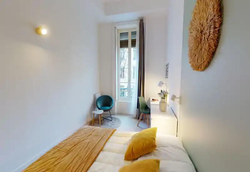Camera da letto luminosa e accogliente con letto ben rifatto, accenti color senape e una finestra che porta luce naturale. Un angolo con poltrona e scrivania vicino alla finestra offre spazio per lavorare o rilassarsi.