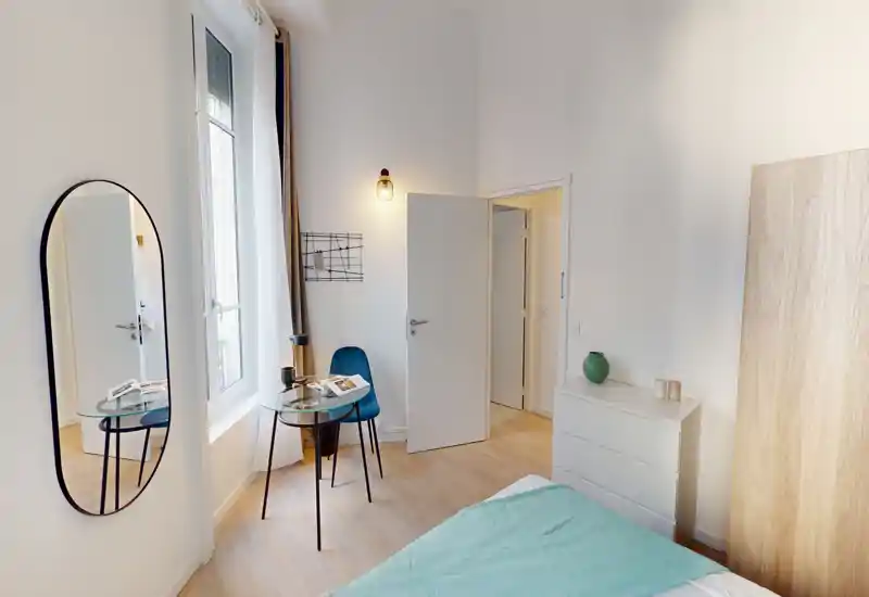 Camera da letto luminosa e minimal con letto visibile e pavimento in legno chiaro. Un piccolo tavolo in vetro con sedia vicino alla finestra, specchio alto e comò per spazio e stile.