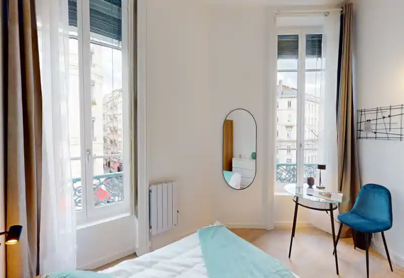 Camera da letto luminosa e moderna con grandi finestre, letto ben rifatto e un piccolo tavolo vicino alla finestra — ambiente arioso con vista sulla città.