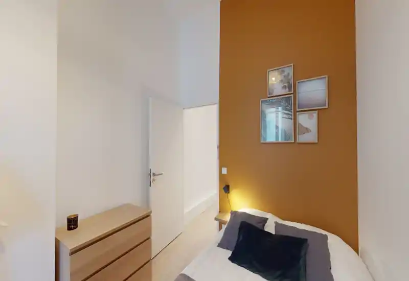 Camera da letto compatta con letto matrimoniale accogliente su una parete color ocra, lampada da comodino e quadri; cassettiera in legno chiaro e tessili morbidi rendono l'ambiente luminoso e invitante.