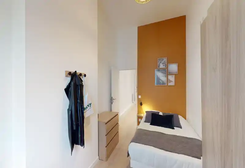 Camera singola compatta e luminosa con letto ben rifatto, parete d'accento calda e lampada da comodino; ideale per un soggiorno confortevole.