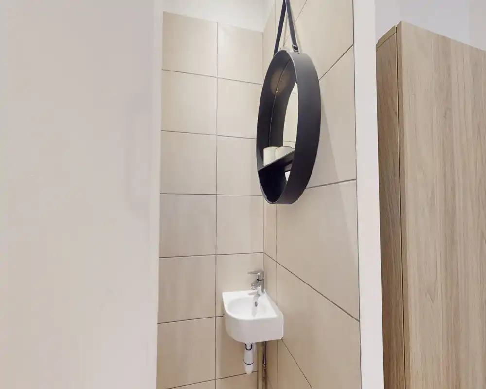 Compact half-bath with a small corner wall-mounted sink and a round hanging mirror, tiled walls and a light wood cabinet visible at the edge — a neat, minimalist guest washroom.