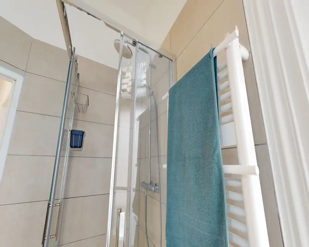 Bagno moderno e luminoso con cabina doccia in vetro, soffione a pioggia e termoarredo bianco con un asciugamano turchese.