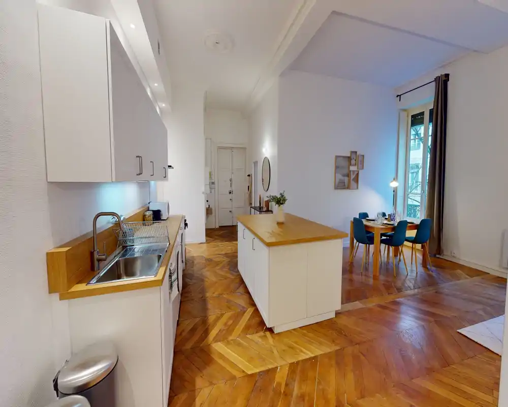 Cucina open-space luminosa con isola centrale e piani in legno che si affacciano su una zona pranzo accogliente; pavimento in parquet caldo e pensili bianchi moderni creano un ambiente invitante e ben illuminato.