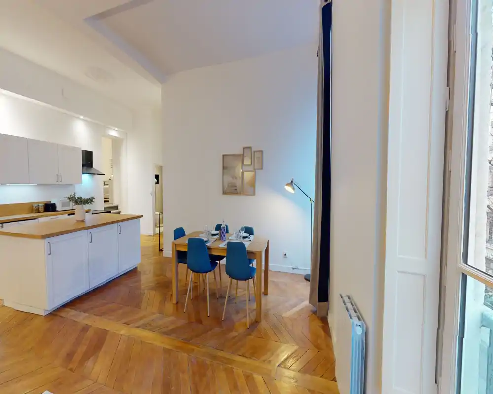 Zona pranzo open-space con tavolo in legno per quattro persone, sedie blu e caldo parquet a spina di pesce; bancone cucina adiacente e ampia finestra che illumina l'ambiente.