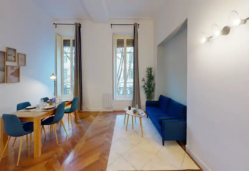 Soggiorno luminoso e di design con divano blu scuro, tappeto chiaro e piccolo tavolino rotondo vicino alle finestre alte; tavolo da pranzo in legno con sedie a sinistra sul pavimento in parquet lucido.