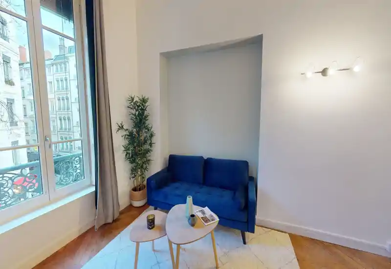 Piccolo soggiorno elegante con divano blu scuro per due persone, tavolini a nido, tappeto e ampia finestra che illumina bene l'ambiente.
