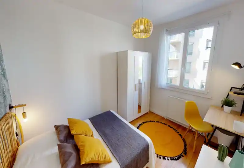 Bright, styled bedroom with a large bed dressed in neutral linens and yellow accent pillows, a wardrobe, desk and window providing abundant natural light.