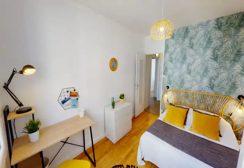 Bright, stylish bedroom with a rattan headboard, leafy accent wallpaper and mustard-yellow cushions; includes a desk and bedside lamps for work and relaxation.