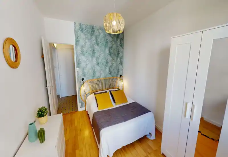 Bright, stylish bedroom with a rattan headboard, patterned accent wall and a comfortable double bed dressed in white linen and mustard cushions. The room features warm wooden floors, a white wardrobe and soft pendant lighting for a cosy atmosphere.