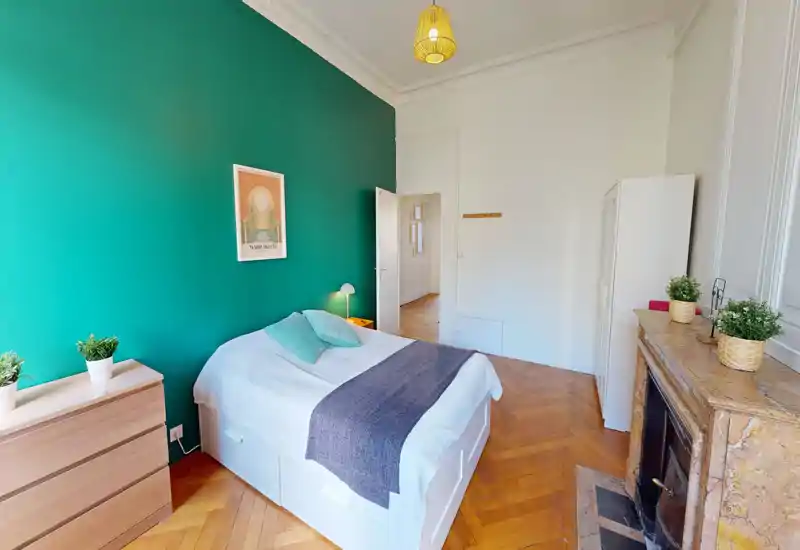 Bright, stylish bedroom with a teal accent wall, double bed dressed in white linens and soft cushions, wooden parquet floor and decorative fireplace — ideal for a cozy rental stay.