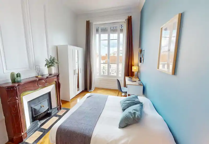 Camera da letto luminosa e di stile con letto matrimoniale, cuscini decorativi e un camino classico. Ampia finestra che illumina la stanza e una piccola scrivania vicino alla finestra per lavorare.