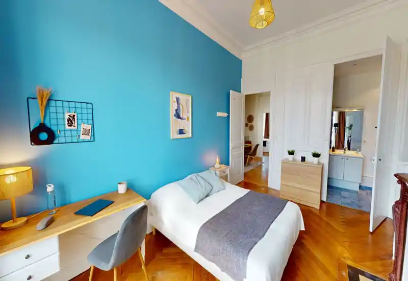 Camera da letto luminosa e di design con letto singolo su una parete blu intensa, scrivania, lampada da comodino e pavimento in parquet — perfetta per un soggiorno accogliente in città.