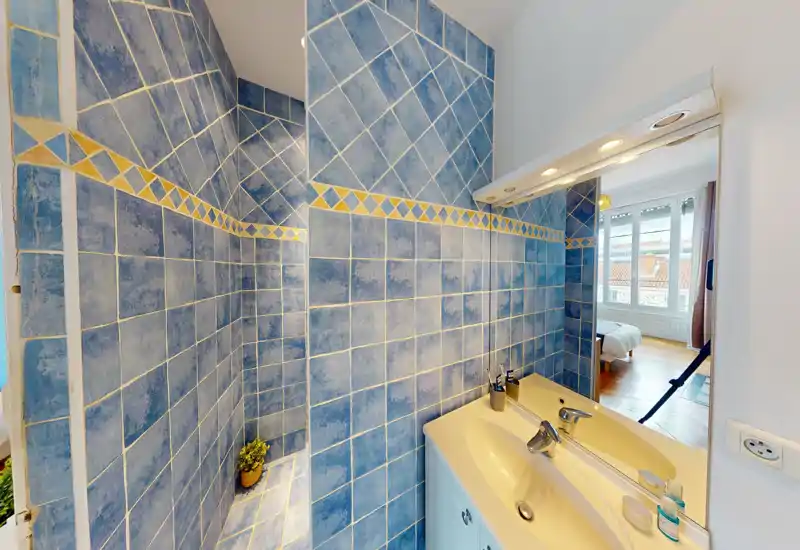 Bagno compatto con pareti piastrellate blu, nicchia doccia integrata e lavabo bianco con specchio; tramite la porta si vede la finestra e la camera da letto.