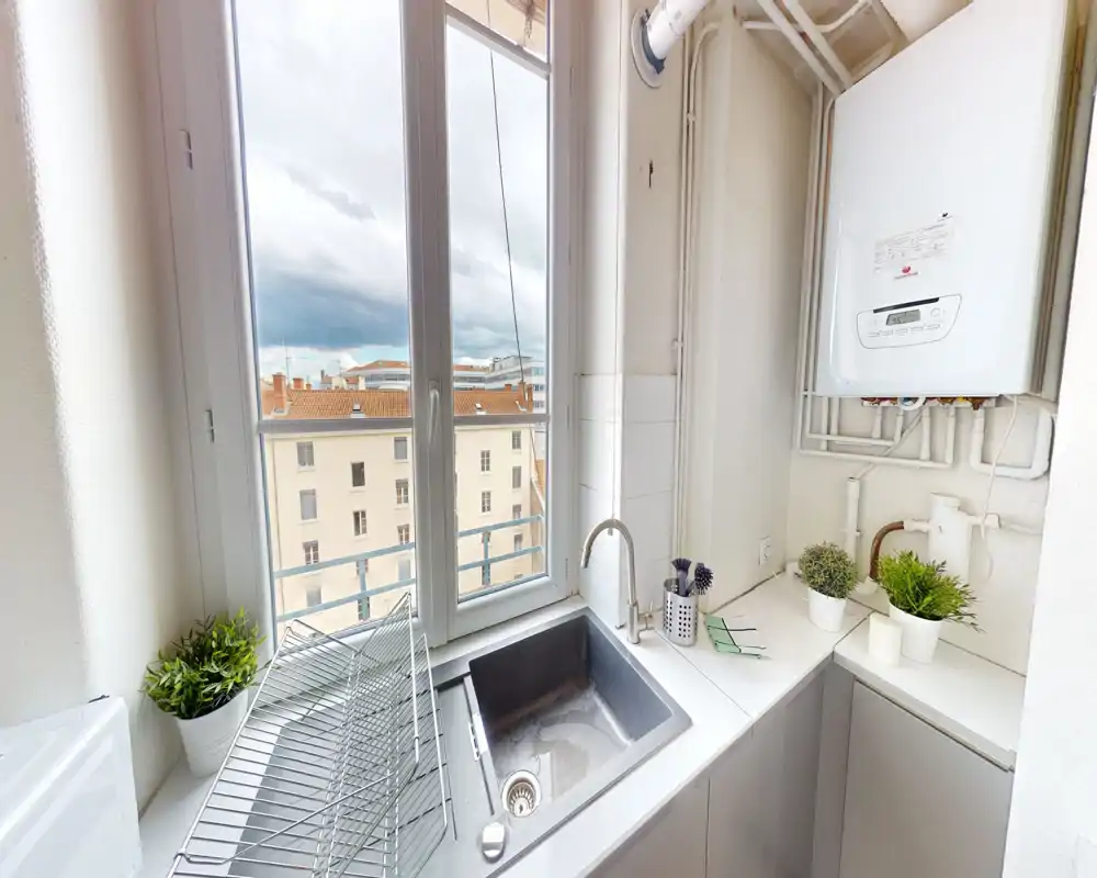 Angolo cucina compatto e luminoso con lavello in acciaio, rubinetto moderno e ampia finestra con vista sulla città; piani di lavoro ordinati con piccole piante e caldaia a vista.