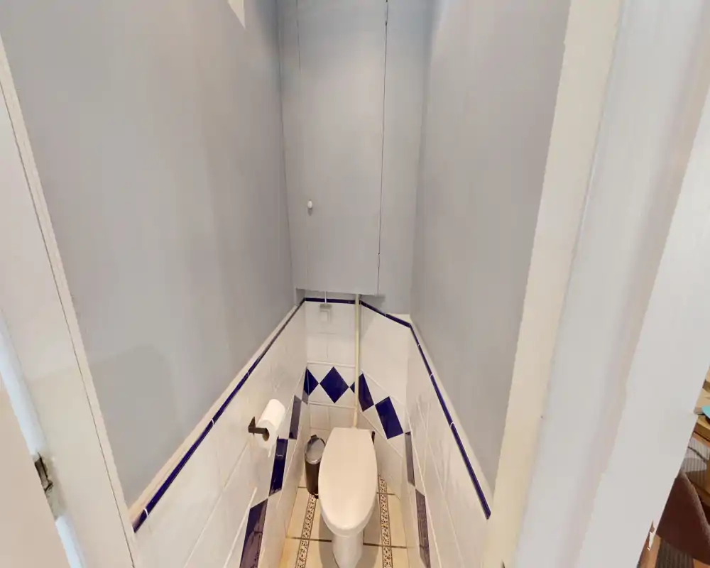 WC compatto con piastrelle bianche e rombi blu, porta rotolo a parete e piccolo cestino — luminoso e disposto in modo efficiente.