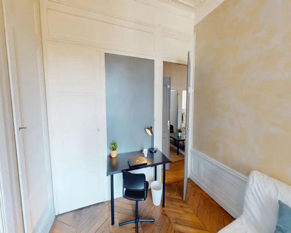 A bright, stylish living area with chevron wood floors, a compact work desk with lamp and chair, and a cozy sofa corner visible at the edge — ideal for a small apartment living room.
