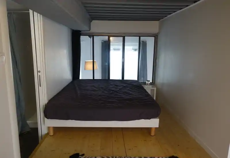 Camera da letto matrimoniale luminosa con pavimento in legno, letto semplice con biancheria scura e comodino con lampada, separata dalla finestra luminosa da una parete vetrata e tende.