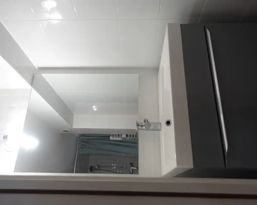 Lavabo moderno di piccolo bagno con specchio e doccia visibile nello specchio; piastrelle bianche pulite e mobile lavabo grigio.
