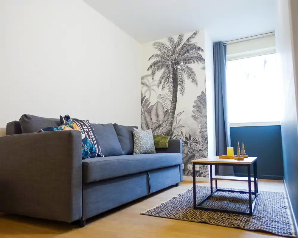 Espace salon compact et élégant avec canapé bleu confortable, coussins à motifs, petite table basse et une fresque murale de palmier — lumière naturelle claire et accueillante.