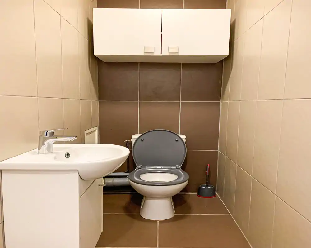 Petite salle de bain propre avec lavabo mural, WC bouchon ouvert et meuble de rangement au-dessus ; carrelage beige et marron offrant un look moderne et compact.