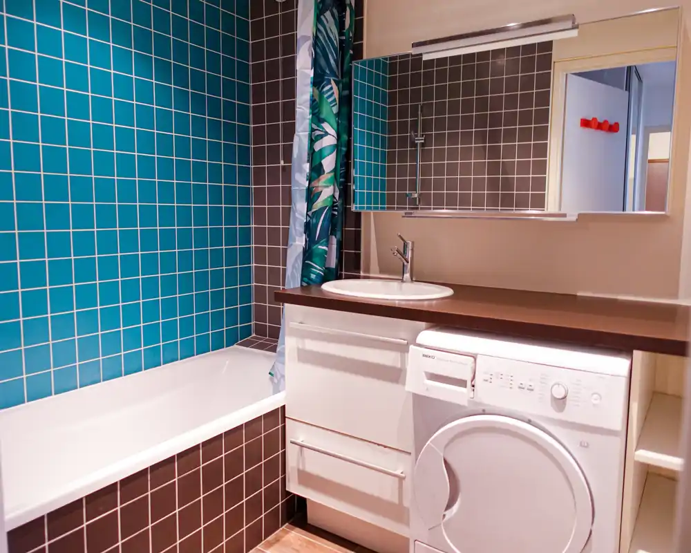Salle de bain moderne avec baignoire, murs carrelés turquoise et brun foncé, lavabo intégré sur plan et lave-linge/sèche-linge à chargement frontal sous le plan de travail.