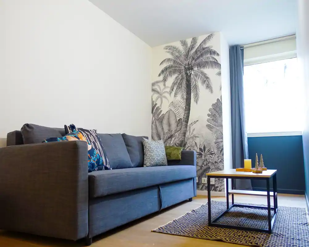 Salon lumineux et élégant avec un canapé gris, des coussins décoratifs, une table basse et une fresque murale tropicale à côté d’une grande fenêtre qui inonde la pièce de lumière naturelle.
