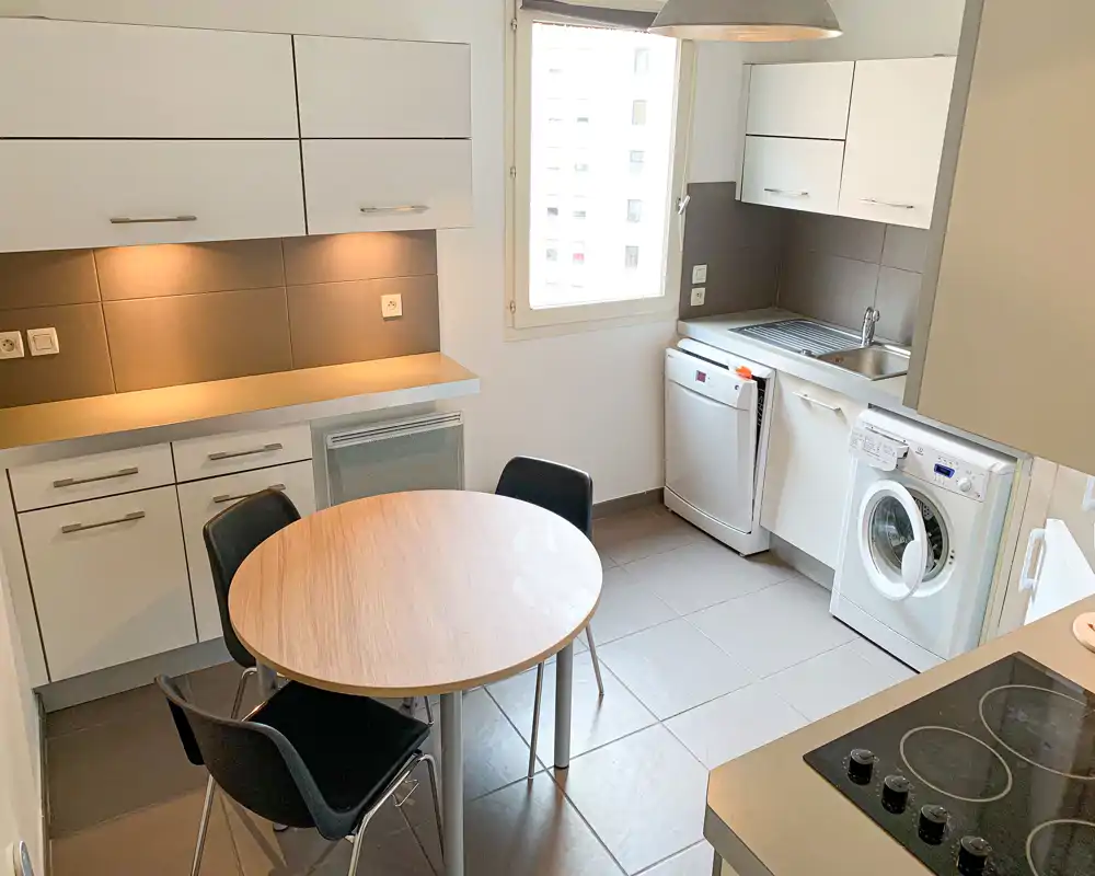 Cuisine moderne et compacte avec électroménagers intégrés et petite table ronde. Espace lumineux avec placards blancs, lave-linge et lave-vaisselle visibles, adapté à un appartement en ville.
