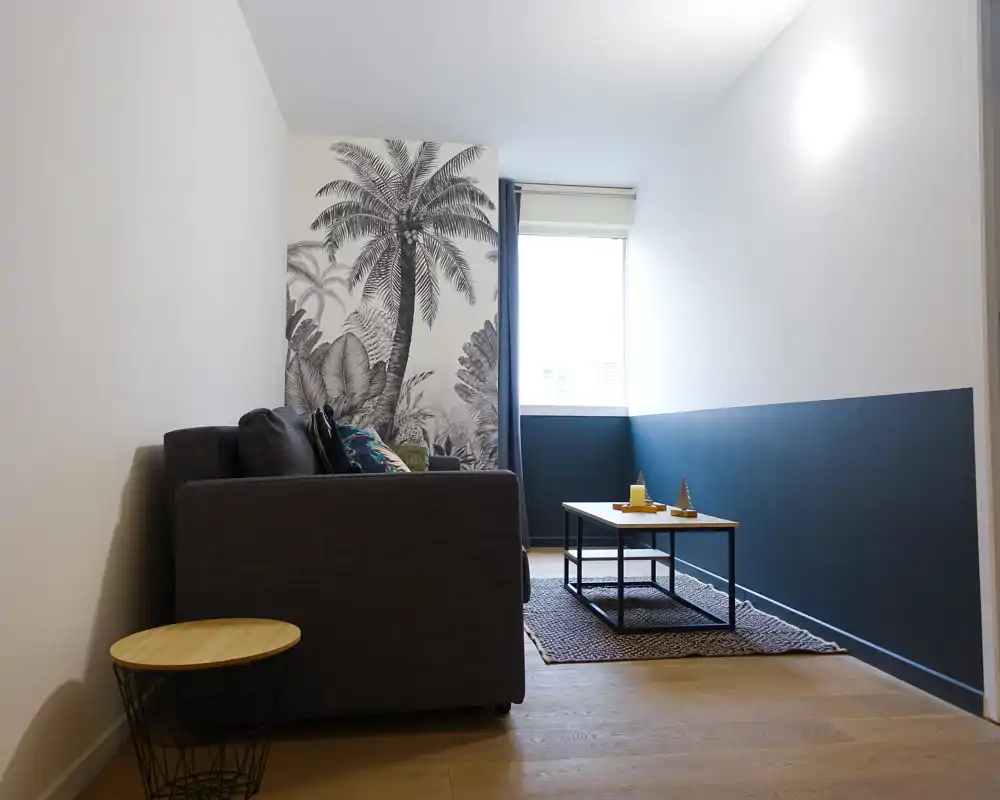 Espace salon compact et élégant avec canapé gris, fresque de palmiers, table basse et lumière naturelle par la fenêtre — idéal pour un coin détente chaleureux.