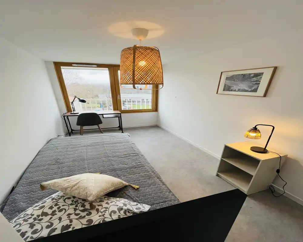 Camera da letto luminosa e minimalista con letto matrimoniale in primo piano, una scrivania vicino alla grande finestra e lampade calde a sospensione e da comodino che creano un'atmosfera accogliente.