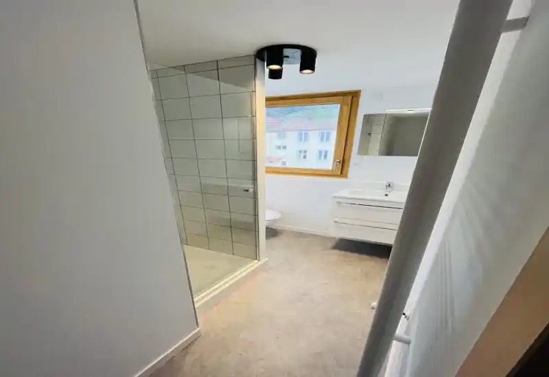 Bright modern bathroom with a glass shower enclosure, wall-mounted vanity with drawers, and a large window providing natural light.