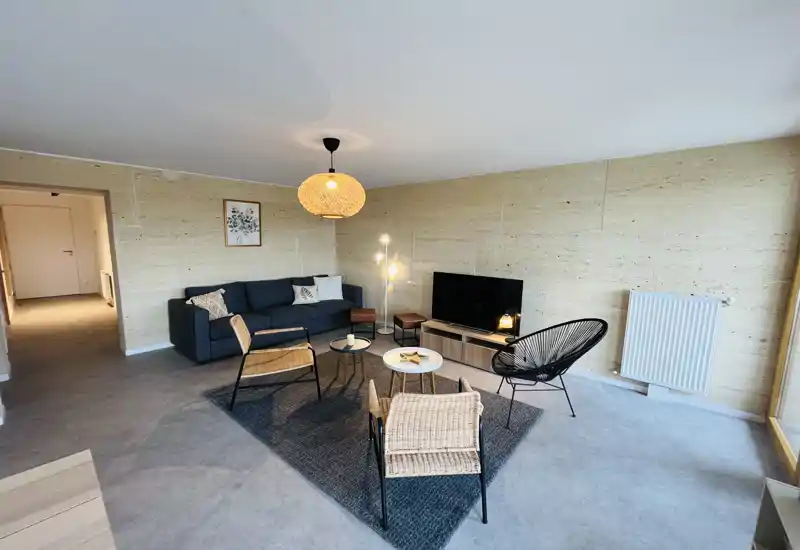 Bright, modern living room with a comfortable sofa, rattan chairs, a central rug and a media console with TV. Warm pendant and floor lamps add cozy ambience; large window (right) provides natural light.