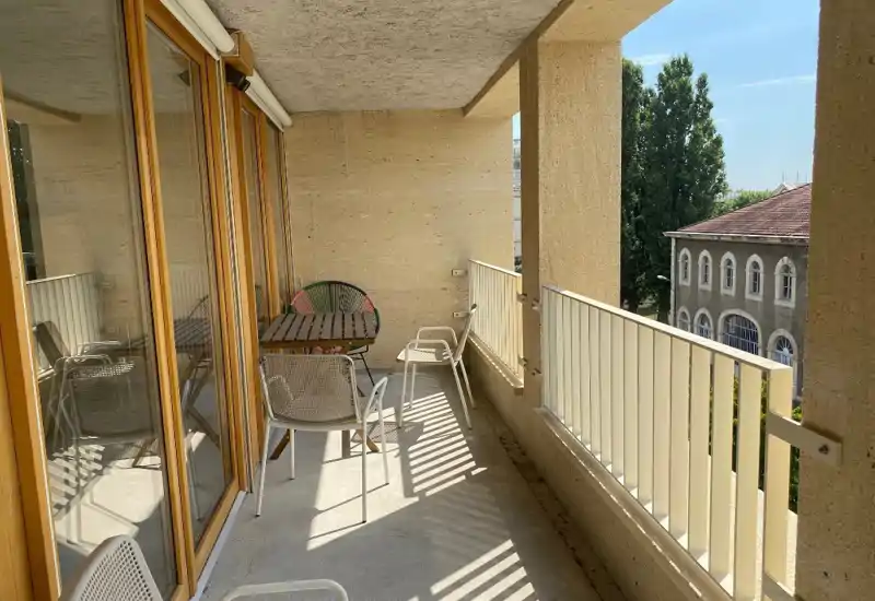 Sunny furnished balcony with a small table and chairs, sheltered by a concrete overhang and offering street-facing views. Ideal for morning coffee or light outdoor dining.
