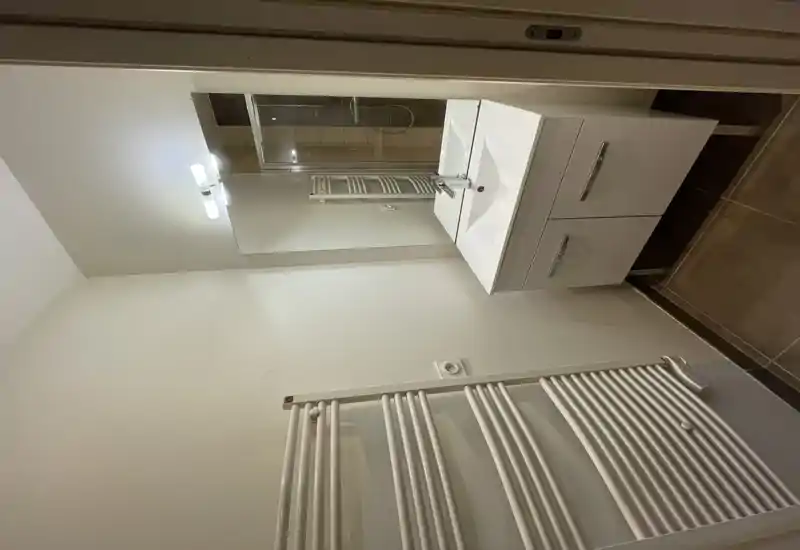 Baño moderno y compacto con mueble lavabo blanco, espejo grande, radiador toallero y vista parcial de una ducha de cristal. Luminoso, limpio y práctico, ideal para un apartamento.
