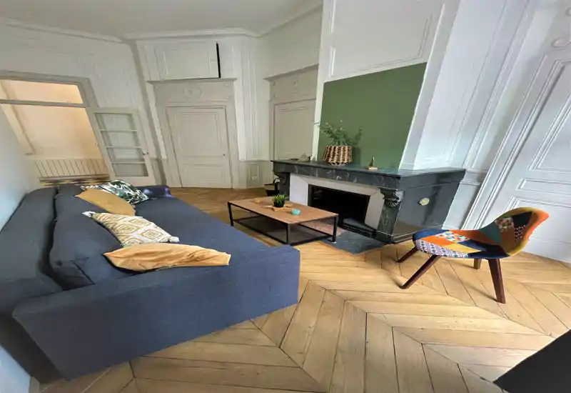 Salón con estilo, gran sofá oscuro, butaca de acento con estampado, mesa de café y chimenea decorativa — espacio luminoso y bien decorado, ideal para relajarse.