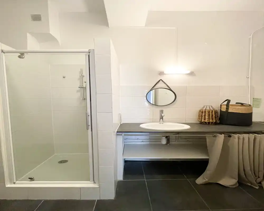 Salle de bain propre et moderne avec une douche carrelée, lavabo intégré dans un plan et un petit miroir rond au-dessus. Tons neutres et rangement pratique sous le lavabo.