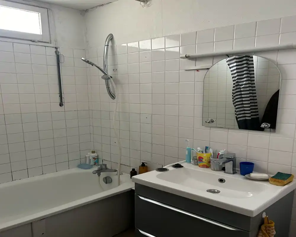 Bagno compatto con pareti piastrellate bianche, vasca con doccia a parete, lavabo con rubinetteria cromata e specchio pensile. Ambiente luminoso con arredi moderni e puliti.
