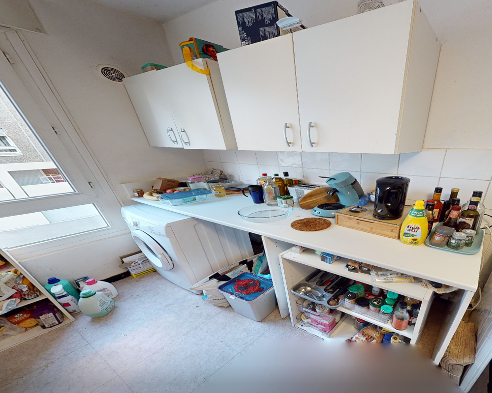 Cucina funzionale e luminosa con pensili, piano di lavoro, piccoli elettrodomestici e lavatrice; un po' disordinata ma pratica per l'uso quotidiano.