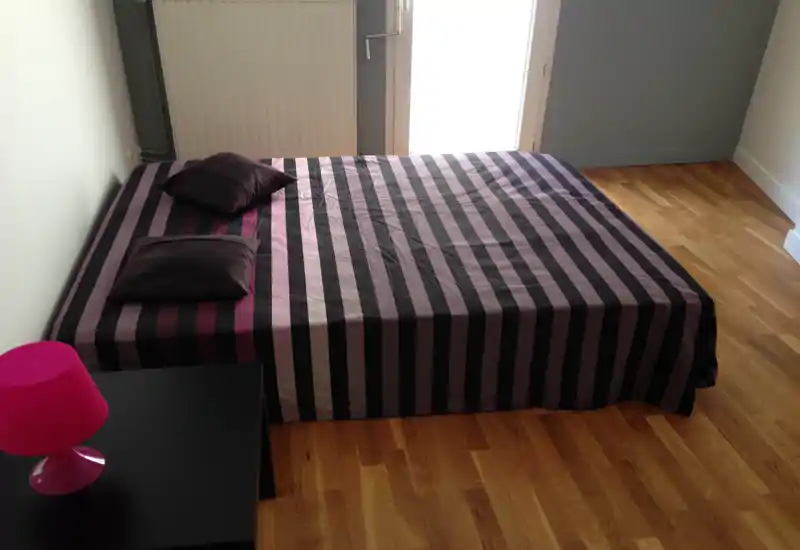 Bright, minimalist bedroom with a low double bed dressed in a striped cover, two pillows and natural light from a door/window. Hardwood floors and simple bedside table with a pink lamp complete the clean, modern look.
