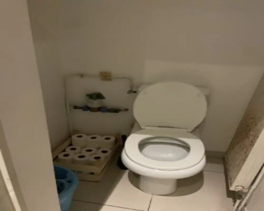 Compact bathroom with a standard toilet, tiled floor and visible plumbing; a stack of toilet paper rolls is stored beside the bowl.